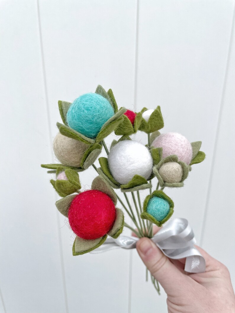 May include: A bouquet of felt flowers with green leaves and colorful felt balls in shades of pink, white, red, and blue. The bouquet is tied with a white ribbon.