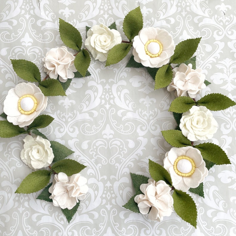 May include: A white felt garland with green leaves and white felt flowers. The flowers have yellow centers.