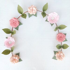 May include: A garland of felt flowers in shades of pink and white, with green felt leaves, is strung together to form a circle. The garland is perfect for adding a touch of spring to any room.