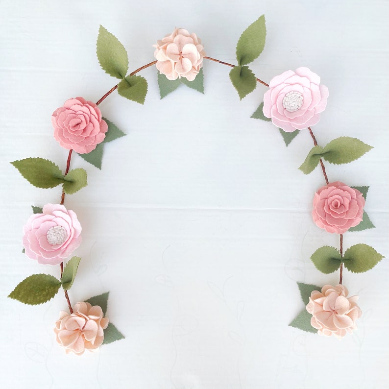 May include: A garland of felt flowers in shades of pink and white, with green felt leaves, is strung together to form a circle. The garland is perfect for adding a touch of spring to any room.