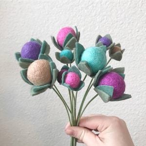 May include: A bouquet of felt flowers with green leaves and colorful felt balls in shades of pink, purple, teal, and beige.