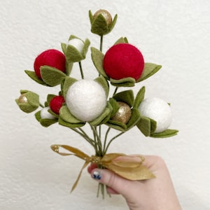 May include: A bouquet of felt Christmas ornaments with red, white, and gold accents. The ornaments are attached to green felt stems and tied together with a gold ribbon.
