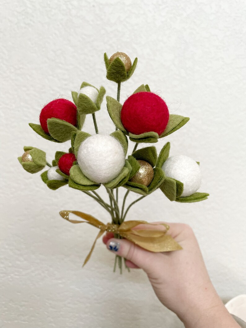 May include: A bouquet of felt Christmas ornaments with red, white, and gold accents. The ornaments are attached to green felt stems and tied together with a gold ribbon.