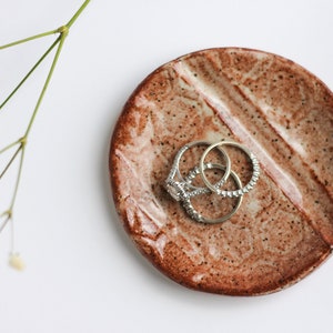 Könnte beinhalten: Eine braune Keramik-Ring Schale mit einem gesprenkelten Überzug. Zwei silberne Ringe liegen in der Schale.