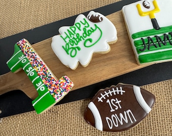May include: Four decorated sugar cookies on a wooden board. One cookie is shaped like the number one with green icing and white sprinkles. Another cookie is shaped like a football with brown icing and white stitching. The third cookie is white with green icing that says "Happy Birthday". The fourth cookie is white with green and black icing that says "Jamie".