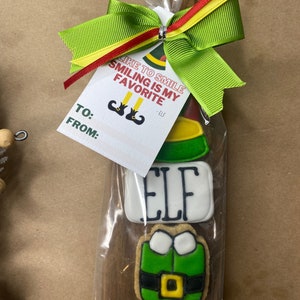 May include: A set of Christmas cookies decorated with elf-themed designs, including a green elf hat, a white cookie with the word "ELF" in black lettering, a green elf with a yellow belt, and a red and white candy cane. The cookies are packaged in a clear plastic bag with a wooden paddle and a green bow. The bag also includes a gift tag with the text "Like to smile, smiling is my favorite - Elf" and space for "To:" and "From:".