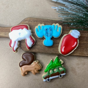 May include: Five Christmas-themed sugar cookies decorated with icing. The cookies are shaped like a moose, a Christmas tree, a squirrel, a stocking, and a Christmas ornament. The cookies are on a wooden cutting board.