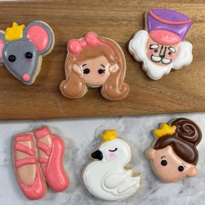 May include: A collection of decorated sugar cookies in the shape of ballet-themed characters. The cookies include a mouse with a crown, a ballerina, a Nutcracker, ballet slippers, a swan, and a girl with a bun. The cookies are decorated with pink, white, gray, and brown icing.