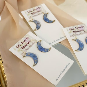May include: A pair of crescent moon earrings with blue pressed flowers. The earrings are handmade in Seattle and are available for purchase on Instagram @ggjewelsblooms.