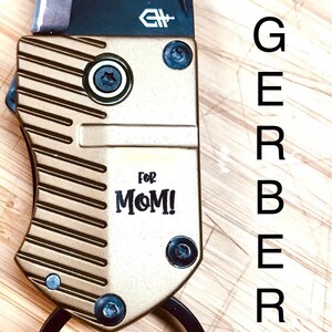 May include: A gold and black pocket knife with a ring on the handle. The knife has a Gerber logo and the words "For Mom!" engraved on the side.