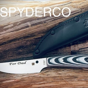 May include: A Spyderco knife with a black and white patterned handle and a stainless steel blade. The blade is engraved with the words "For Dad" and a spider logo. The knife comes with a black leather sheath.