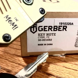 May include: A beige Gerber Key Note CYBR, FE 30-001692 key chain with the words "I love MOM!" printed on it. The key chain is packaged in a brown box with a white label that says "GERBER KEY NOTE CYBR, FE 30-001692 MADE IN CHINA" and the product number 1910320A.