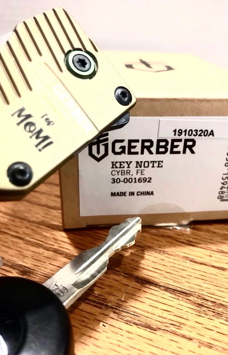 May include: A beige Gerber Key Note CYBR, FE 30-001692 key chain with the words "I love MOM!" printed on it. The key chain is packaged in a brown box with a white label that says "GERBER KEY NOTE CYBR, FE 30-001692 MADE IN CHINA" and the product number 1910320A.