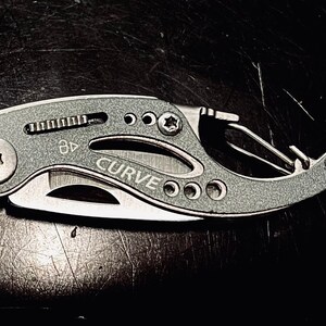 K&ouml;nnte beinhalten: Ein silbernes Multitool mit schwarzem Griff und einer gebogenen Klinge. Das Werkzeug hat einen kleinen Karabinerhaken und einen kleinen Schraubendreher. Das Werkzeug ist mit "CURVE" beschriftet.