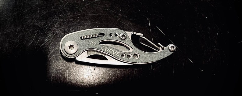 K&ouml;nnte beinhalten: Ein silbernes Multitool mit schwarzem Griff und einer gebogenen Klinge. Das Werkzeug hat einen kleinen Karabinerhaken und einen kleinen Schraubendreher. Das Werkzeug ist mit "CURVE" beschriftet.