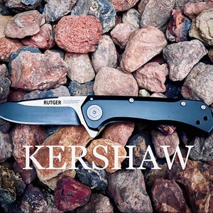 Könnte beinhalten: Ein schwarzes Kershaw Taschenmesser mit einer silbernen Klinge und den Worten "RUTGER" und "Inox" auf dem Griff. Das Messer liegt auf einem Bett aus kleinen roten und grauen Steinen.