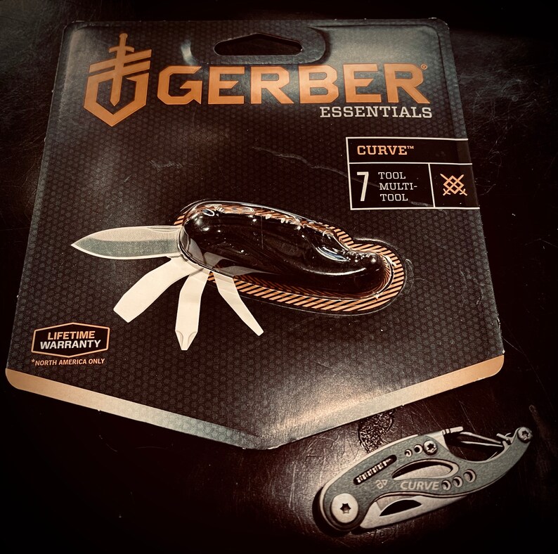 K&ouml;nnte beinhalten: Ein schwarzes Gerber Essentials Curve 7-teiliges Multitool mit einer lebenslangen Garantie in Nordamerika. Das Multitool ist ge&ouml;ffnet und die Klinge ist sichtbar. Die Verpackung ist schwarz mit goldenen Schriftz&uuml;gen und einem Wabenmuster.