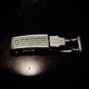 K&ouml;nnte beinhalten: Ein silbernes und graues Multitool mit einem schwarzen Griff. Der Griff tr&auml;gt die Aufschrift "GERBER" in Pr&auml;gung.