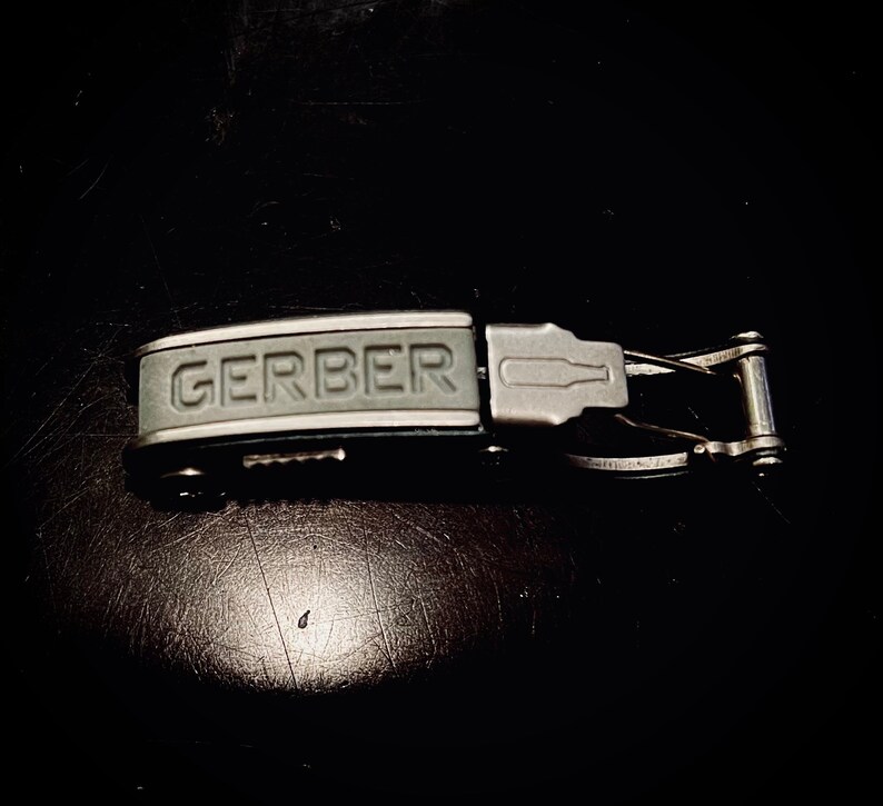 K&ouml;nnte beinhalten: Ein silbernes und graues Multitool mit einem schwarzen Griff. Der Griff tr&auml;gt die Aufschrift "GERBER" in Pr&auml;gung.