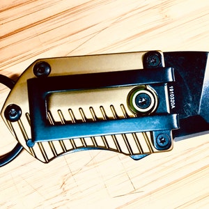 May include: A gold and black keychain utility knife with a ring and a blade. The blade is partially extended and the knife has a textured surface. The knife has the number 1910320A printed on it.