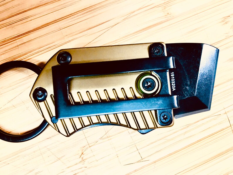 May include: A gold and black keychain utility knife with a ring and a blade. The blade is partially extended and the knife has a textured surface. The knife has the number 1910320A printed on it.