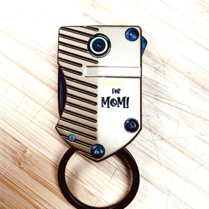 May include: A gold-coloured metal keychain with a black ring. The keychain has a textured surface and the words "for MOM!" printed on it.