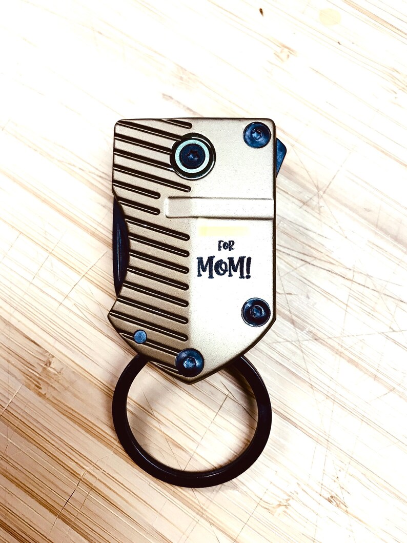 May include: A gold-coloured metal keychain with a black ring. The keychain has a textured surface and the words "for MOM!" printed on it.