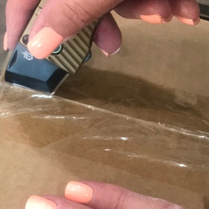 May include: A hand holding a small, silver and brown pocket knife with a black handle. The knife is being used to cut through clear plastic tape on a brown cardboard box. The hand has peach-colored nail polish with white and black designs.