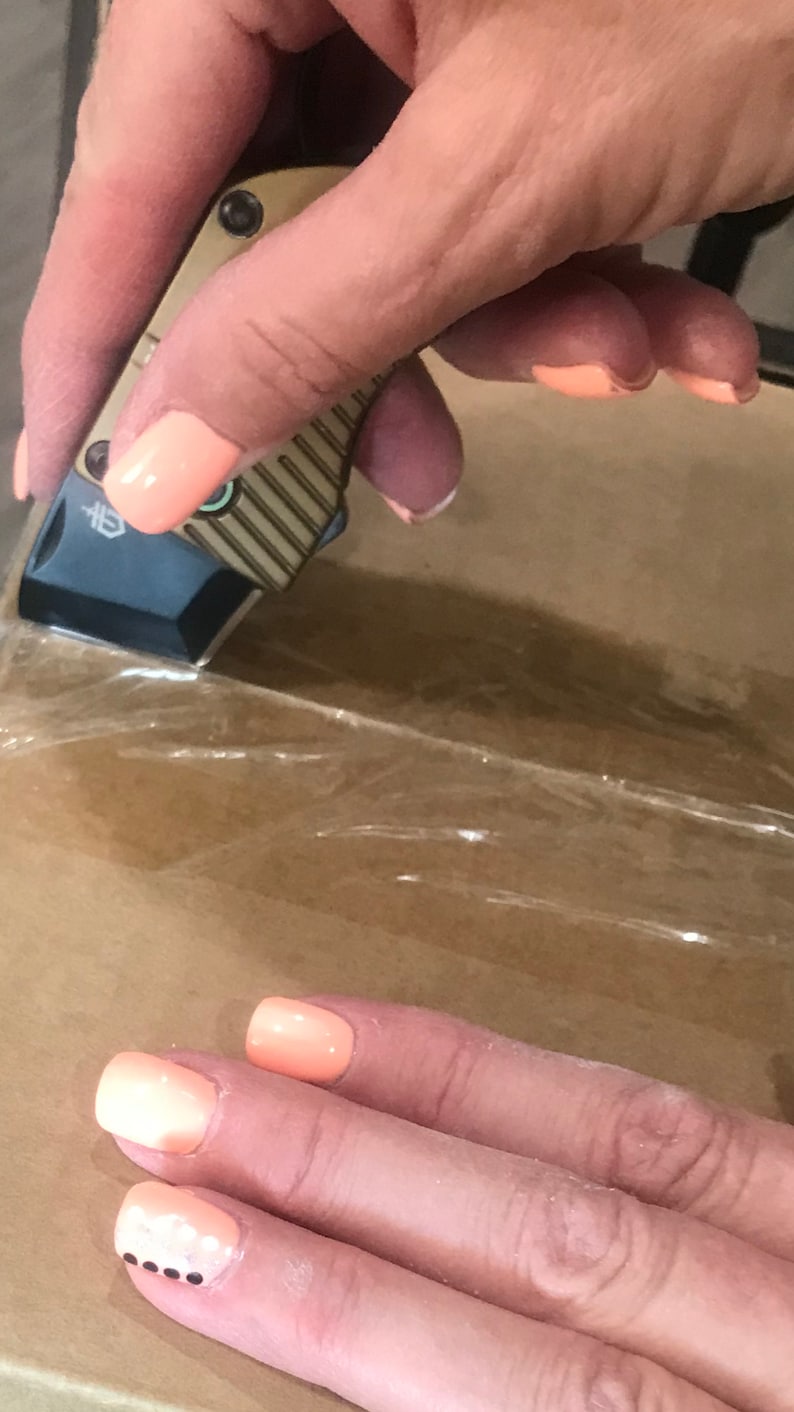 May include: A hand holding a small, silver and brown pocket knife with a black handle. The knife is being used to cut through clear plastic tape on a brown cardboard box. The hand has peach-colored nail polish with white and black designs.