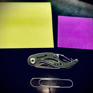 K&ouml;nnte beinhalten: Ein schwarzes Multitool mit dem Wort "CURVE" darauf, ein gelber Haftnotiz, ein lila Haftnotiz und eine silberne B&uuml;roklammer auf einer schwarzen Oberfl&auml;che.