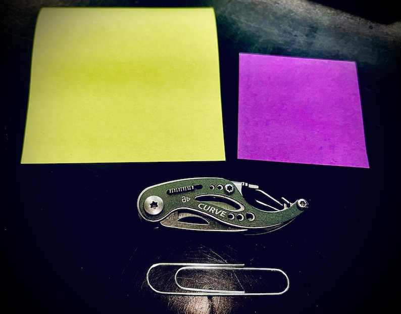 K&ouml;nnte beinhalten: Ein schwarzes Multitool mit dem Wort "CURVE" darauf, ein gelber Haftnotiz, ein lila Haftnotiz und eine silberne B&uuml;roklammer auf einer schwarzen Oberfl&auml;che.
