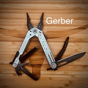 May include: A silver and black Gerber multi-tool, with the brand name visible. The tool includes pliers, a knife, and other implements. The tool is open, showing its functions. It rests on a light brown wooden surface.