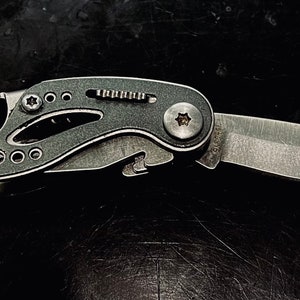 K&ouml;nnte beinhalten: Ein schwarzes Multifunktions-Taschenmesser mit einer silbernen Klinge und einer Vielzahl von Werkzeugen, darunter ein Flaschen&ouml;ffner, ein Schraubendreher und eine Feile.