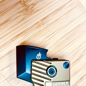 May include: A blue and gold multi-tool with a Gerber logo. The tool has a textured surface and a black screw.