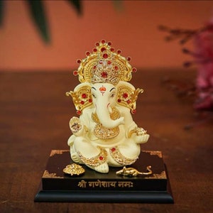 Peut inclure: Statue ivoire du dieu hindou Ganesha, ornée d'accents dorés et rouges. La statue présente une couronne et des bijoux détaillés, assise sur une base noire avec des ornements et du texte dorés. Une petite souris dorée est à la base.
