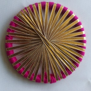 May include: Circular decorative item with a vibrant fuchsia border and gold metallic ribbon radiating from the centre. The design features a woven pattern, creating a starburst effect. The item is set against a plain white background.