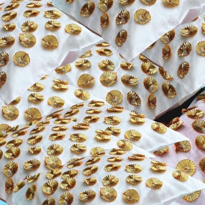 May include: Close-up of several white fabric pieces adorned with gold, circular embellishments. The gold accents are arranged in a scattered pattern across the fabric, creating a textured and decorative effect. The fabric appears to be soft and lightweight.