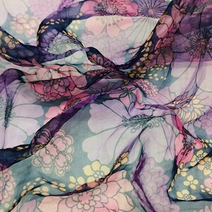 Puede incluir: Una tela transparente con un estampado floral en tonos rosa, morado y amarillo sobre un fondo azul claro. La tela está drapeada, creando pliegues y sombras que realzan el diseño. Las flores son grandes y estilizadas.