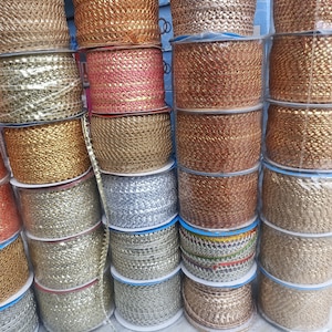 May include: A display of spools of decorative trim in various metallic colors. The trim has a woven or braided texture and is wound on plastic spools. Colors include gold, silver, copper, and pink.