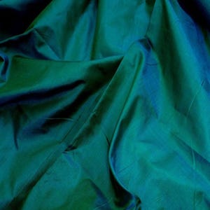 May include: Close-up of a fabric with a teal and blue iridescent sheen. The fabric is heavily draped, creating shadows and highlights that emphasize its texture and color variations. The material appears to be silk or a similar luxurious textile.