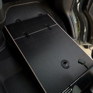May include: A black, rectangular vehicle console tray with a honeycomb pattern. The tray is installed in a vehicle, with a black handle and a small black latch. The tray is designed to fit in the center console area.