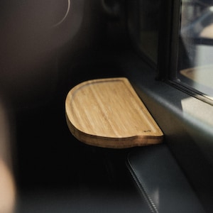 May include: A semi-circular wooden tray with a light brown grain pattern and a raised edge, featuring a small logo mark on the right side. The tray is positioned horizontally against a dark interior surface near a window frame.