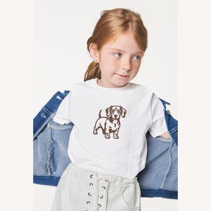 May include: A white t-shirt featuring a brown dachshund dog graphic. The t-shirt is worn by a person with red hair and freckles, paired with white shorts and a denim jacket.