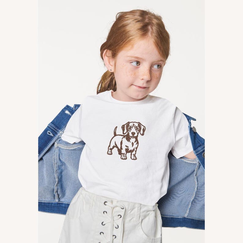 May include: A white t-shirt featuring a brown dachshund dog graphic. The t-shirt is worn by a person with red hair and freckles, paired with white shorts and a denim jacket.