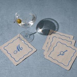 Peut inclure: Un verre à martini transparent avec une olive est posé au-dessus de quatre dessous de verre carrés beiges avec des bords festonnés bleus. Un dessous de verre porte la lettre "M", et un autre une illustration d'olive. Les articles sont sur une surface bleue.