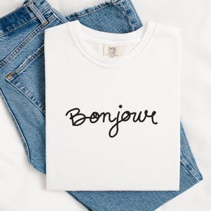 Puede incluir: Una camiseta blanca con la palabra "Bonjour" bordada en escritura negra. La camiseta está doblada y descansa sobre un par de vaqueros azules. La camiseta tiene una etiqueta Comfort Colors.