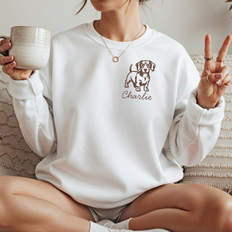 May include: White sweatshirt featuring a brown dachshund dog graphic and the name "Charlie" in a script font. The sweatshirt has a crew neck and long sleeves. A simple gold necklace is visible.