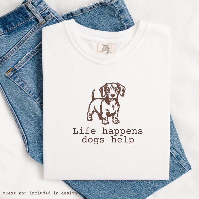 May include: White t-shirt with a brown dachshund graphic and the text "Life happens dogs help." The shirt is folded on top of blue denim jeans. The shirt has a Comfort Colors tag.