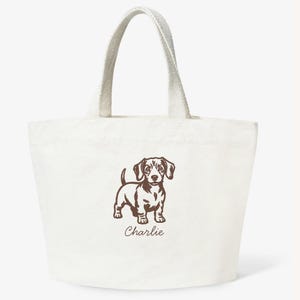 May include: Off-white canvas tote bag with a brown illustration of a dachshund dog. The dog is standing with its head up and the name "Charlie" is printed below the dog. The bag has two handles.
