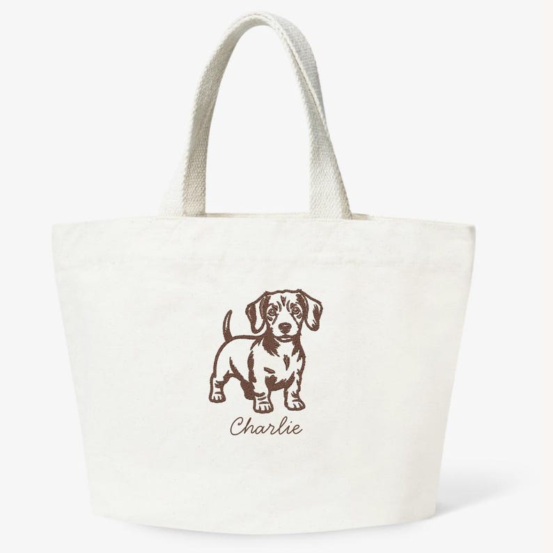 May include: Off-white canvas tote bag with a brown illustration of a dachshund dog. The dog is standing with its head up and the name "Charlie" is printed below the dog. The bag has two handles.